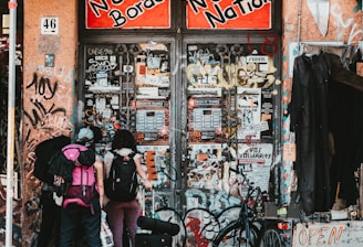 A group of three people with backpacks and luggage stand in front of a graffiti-covered building entrance. The sign above reads 'No Borders No Nation' in bold red and black lettering. Various posters and stickers cover the walls and doors. A bicycle is parked nearby, and a sign with 'OPEN' is visible on the right.