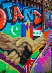 A colorful mural on a brick wall featuring large text in bold graffiti style reading 'Stand with Us'. The artwork includes different flags, a crescent moon, and a star, along with a diverse range of patterns and symbols. There are two hands shaking prominently in the center, emphasizing unity and solidarity.