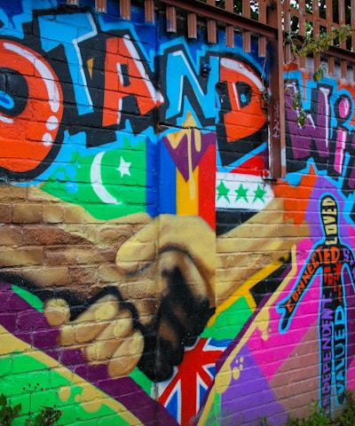 A colorful mural on a brick wall featuring large text in bold graffiti style reading 'Stand with Us'. The artwork includes different flags, a crescent moon, and a star, along with a diverse range of patterns and symbols. There are two hands shaking prominently in the center, emphasizing unity and solidarity.