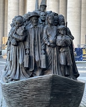 A bronze sculpture depicting a diverse group of people standing closely together on a boat, with expressions and attire representing various cultures and eras. The figures include men, women, and children, some holding luggage, symbolizing themes of migration and unity.