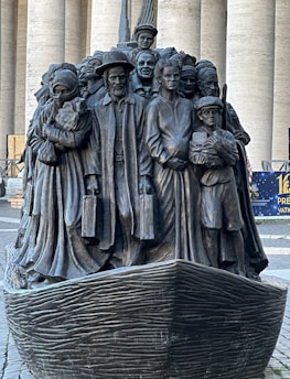 A bronze sculpture depicting a diverse group of people standing closely together on a boat, with expressions and attire representing various cultures and eras. The figures include men, women, and children, some holding luggage, symbolizing themes of migration and unity.